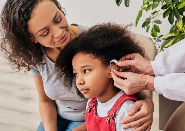 Une enfant dans les bras de sa maman est en train de se faire poser des appareils auditifs par un médecin